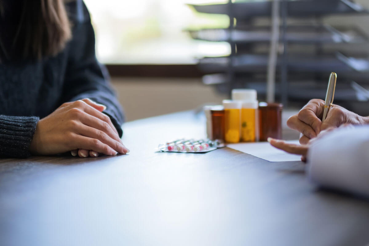 A patient sitting across a table from a doctor, with medication bottles and a prescription pad in view.