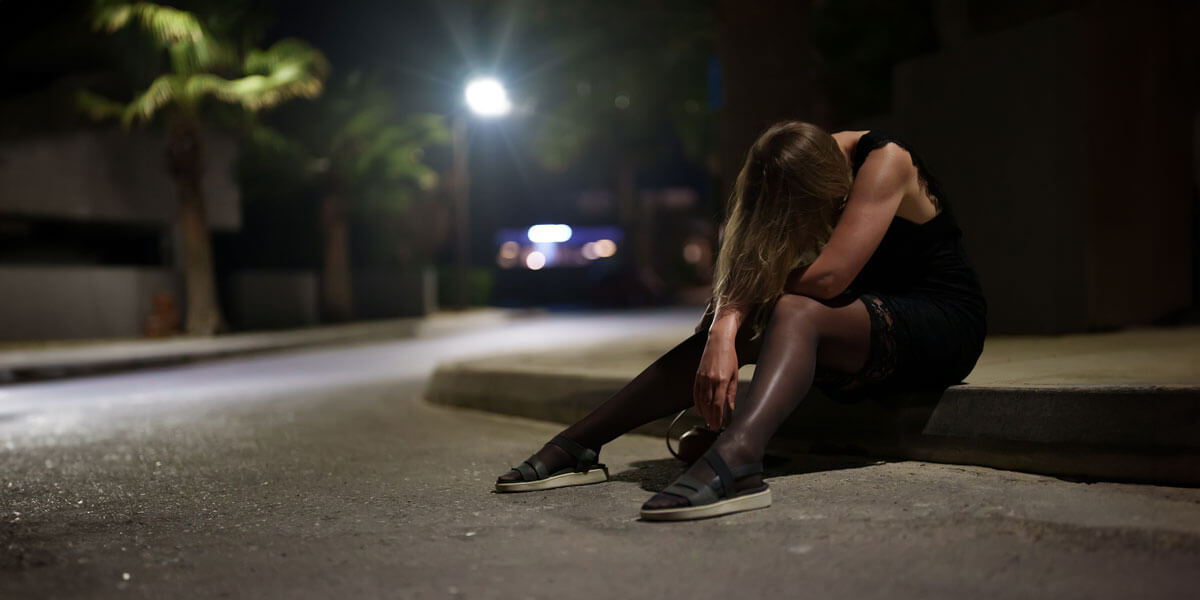 A woman sits on the curb at night, illuminated by streetlights, reflecting on her surroundings in a quiet moment.